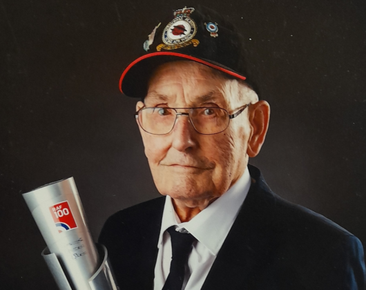 Fred Hooker, an older man in a suit, wears his war medals and displays an RAF 100 trophy with a proud expression.