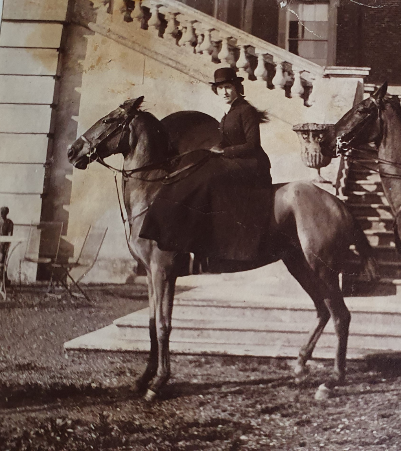 A person in formal attire rides a horse beside elegant stone stairs. The sepia tone gives a historic, timeless feel. The setting is grand and stately.