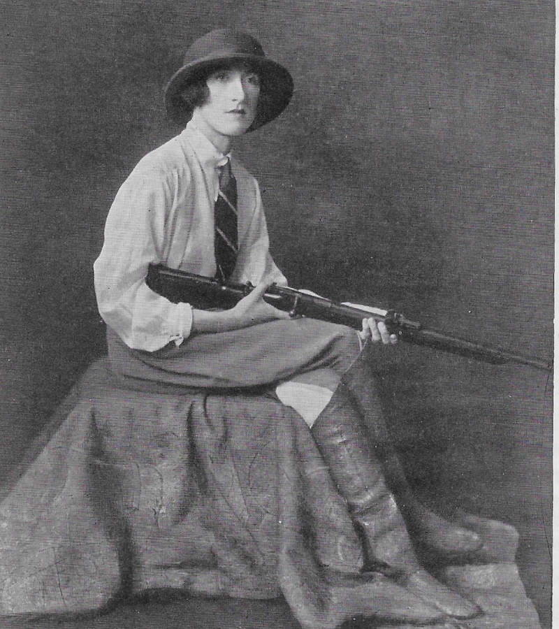 Black and white photo of a person seated with a rifle across their lap. They're wearing a hat, tie, and knee-high boots, conveying a poised, vintage elegance.