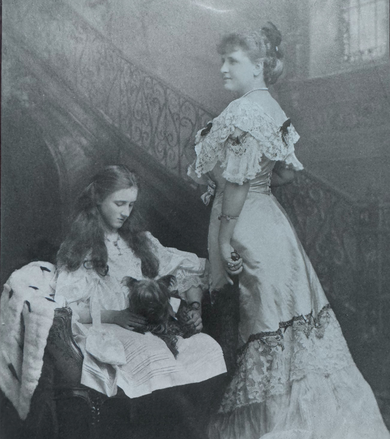 Two women in vintage dresses, one seated holding a small dog, the other standing beside her. They are in front of an ornate staircase, evoking an elegant, nostalgic mood.